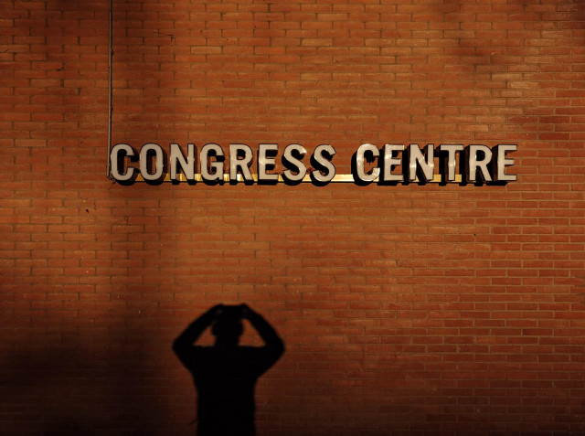 A photo showing red brick wall bathed in warm sunlight with  "Congress Centre" in bold, white letters, and a shadow of a person with their hands raised.