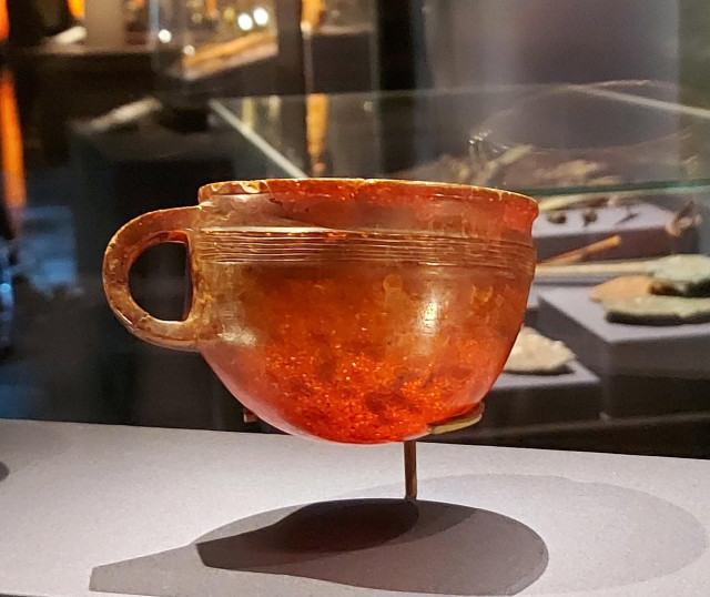 A small amber cup with a single handle displayed in a museum exhibit. The cup features  warm reddish-orange tones and fine horizontal grooves around the upper rim. Light shining through the amber highlights its natural patterns and rich glow.