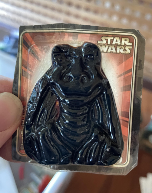 A close up photo of a Star Wars branded lolly packet. Inside its (evidently broken seal) is a lolly moulded in the shape of Jar Jar Binks’ smiling bust. Once multicoloured, it is now an unhealthy deep black.