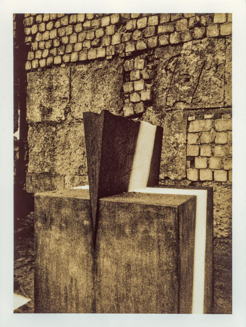 Lith print on Fomatone paper. A steel wedge driven vertically into a freestanding concrete cube, which stands independently in front of a crumbling stone wall. The cube occupies the lower foreground, solid and self-contained. The warm sepia tones of the lith process deepen the textures of metal, concrete, and weathered masonry behind.