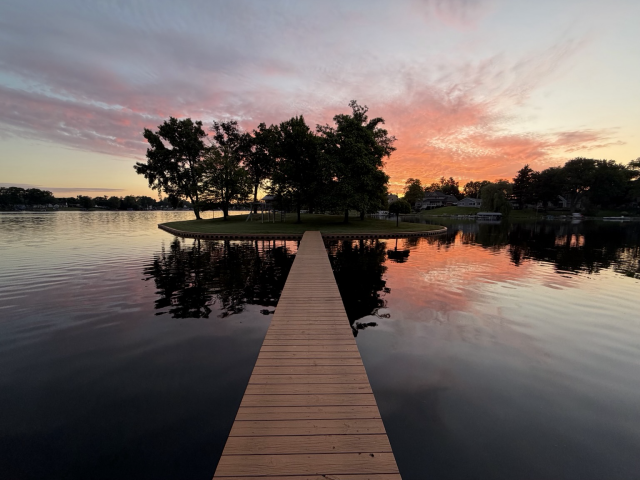 Sunrise photo of pink and peach clouds behind a small island on a reflective lake with a narrow, brown wooden bridge reaching out from the bottom middle to the island.