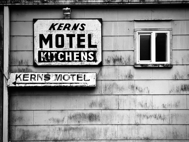 The side of a weathered clapboard motel with one small window, a sign that says “Kerns Motel Kitchens” and another sign that points the way to the main entrance. 
