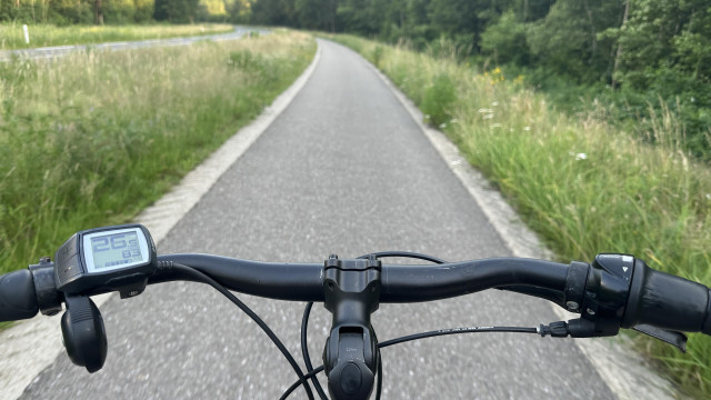 View from a bicycle on a paved trail surrounded by green grass and trees. The bike's handlebar and digital display are visible.