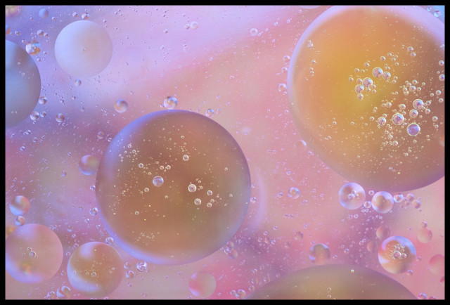 Large and small bubbles of oil float underneath the surface of a Pyrex baking pan of water.  The colors from a blue and pink and gold colored cloth underneath the baking pan shine through the emulsion.