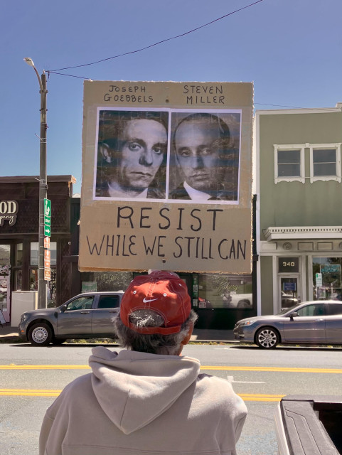 A man stands at the side of a business street holding up a sign that says Resist While We Still Can under side-by-side pictures of Joseph Goebbels and Steven Miller of the Trump regime.