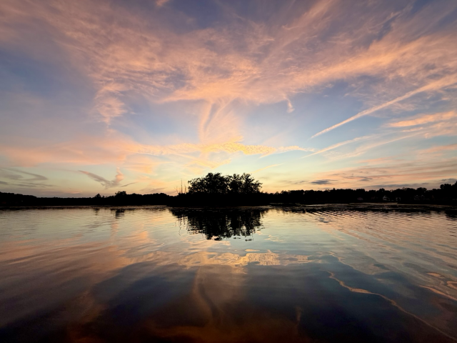 Sunset photograph of wispy pink clouds framing a small island on a reflective lake.