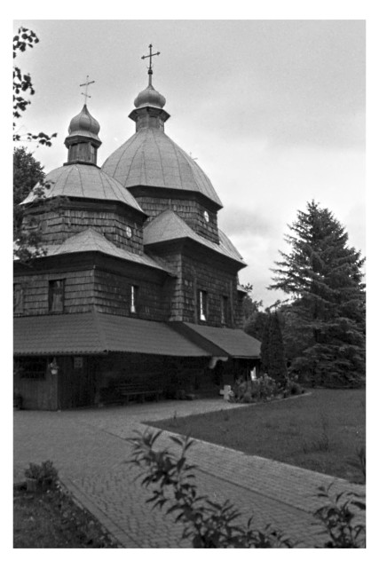Black-and-white photo of a wooden shingled church with two onion domes and crosses against an overcast sky. A tall spruce rises on the right; a paved path and flowerbed lead to the entrance, where a small cross stands among the blooms. 