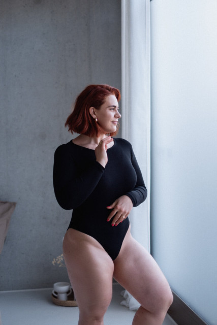 A person with vibrant red, shoulder-length hair is captured in a thoughtful pose. They are wearing a black long-sleeve bodysuit and are crouching slightly, with one hand resting on their chin and the other on their knee. The setting appears to be a minimalist indoor space with a large window on the right, allowing soft natural light to illuminate the scene. The background features a plain, light gray wall, and the floor is light-colored. In the background, there is a small woven basket, a white candle, and a hint of a pink fabric or garment, adding subtle warmth to the otherwise neutral tones of the room.