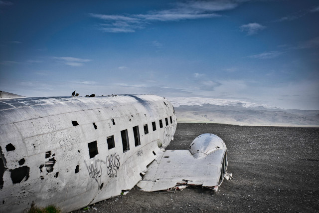 Plane wreckage (US Navy DC-3 Wreckage) in Iceland. An abandoned wreckage on black sand beach, the metal frame all exposed, wings broken, and since aged by the years it's been left here. Some graffiti mark the wreckage, and in the horizon is a glacier rising up towards the mostly clear blue sky.