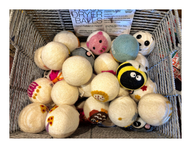 Photo looking down into a wicker basket containing a pile of wool dryer balls, many with designs depicting the faces or entire bodies of animals. The standout is a ball that looks like a confused bumblebee, with black and yellow stripes, white wings, and cross-eyed googly eyes. A paper sign attached to the basket’s interior reads “Wool Dryer Balls - Hand Felted in Nepal” with more information and instructions.