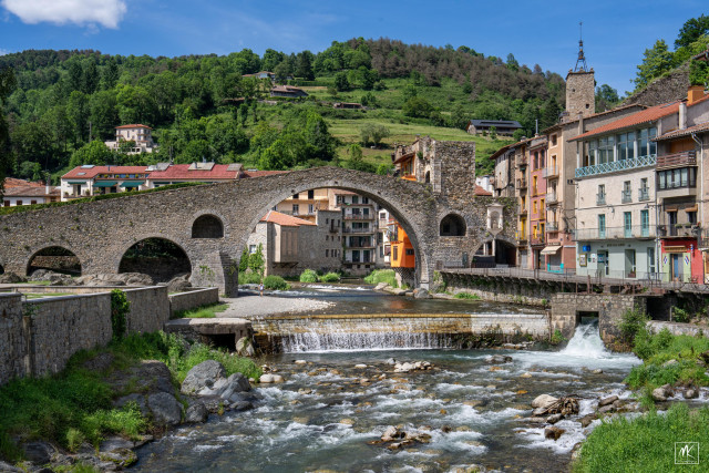 Color photo of an old stone bridge with a large high circular central arch crossing a shallow, stony river with a waterfall over a weir just below it in a Catalonian mountain town.