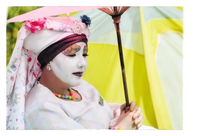 A quiet portrait of a Sister of Perpetual Indulgence, in full white habit and makeup, leaning back on a lawn chair in Dolores Park. They are holding a pink parasol over their head... looking peacefully across the park lawn on a pleasant Sunday afternoon.
