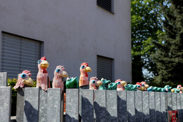 A row of colourful plastic chicken and frog figurines, painted in vibrant pinks, greens, and yellows, perched on top of a grey metal fence in Brno, with a modern building and trees in the background.
