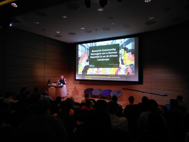 A photo of Cass standing at a podium at the front of a lecture theatre, with the title slide of the presentation on a screen behind them
