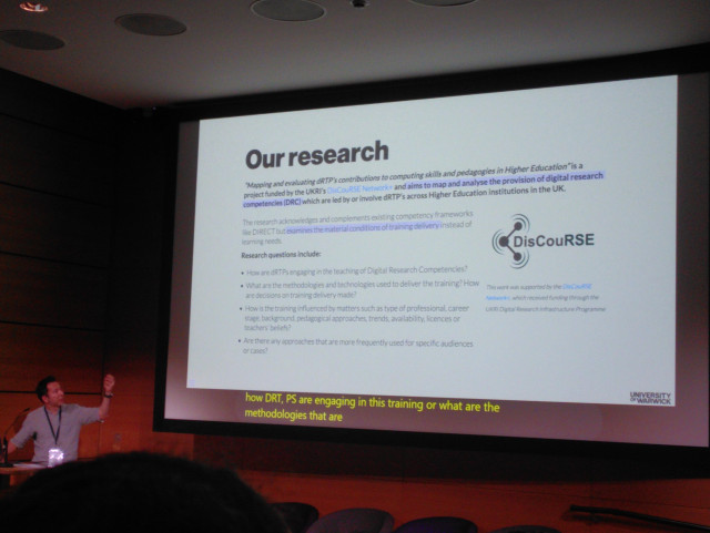 A photo of Carlos presenting a slide with the title "Our Research" 