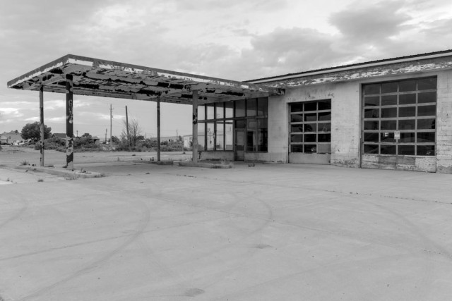 A black and white landscape photo of an old abandoned gas station. All there is is the building and the awning over the fuel pump island. There is nothing else in the photo other than a few weeds growing out of cracks in the pavement. The awning extends out toward the left and the station's garage bays toward the right. The paint on the building is very chipped and flaking off the walls and awning. The sky is cloudy. 