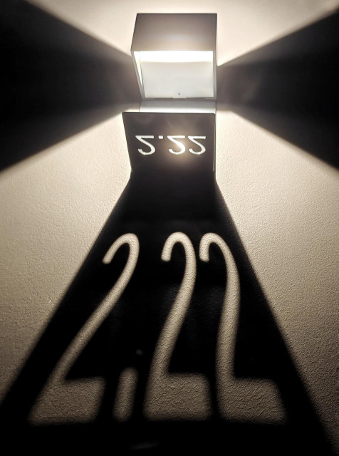 A photo of a wall-mounted light fixture casting a shadow of the numbers 2.22 onto a wall.