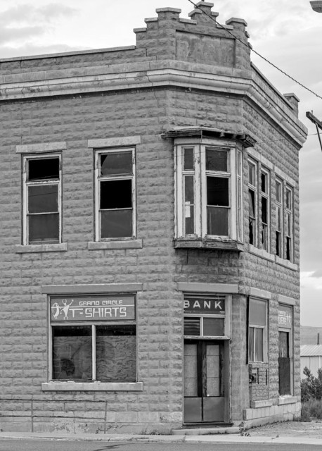A back and white two-story brick building that was once a bank and later a t-shirt shop. The view is looking at the left side of the building with the front door at a 45 degree angle in the corner of the building. We see the less of the right side wall as the camera is pointed more at the left side and front door. The building is made of light colored brick. Over the front door is a glass pane that says, "Bank," across the top. On the left wall is a large window and across the top it says, "Grand Circle T-shirts," with images of ancient petroglyphs in front of the writing. Above the single window on the second floor are two long narrow windows. On the second floor above the front door is a bay window. Windows can also be seen on the right side wall. 