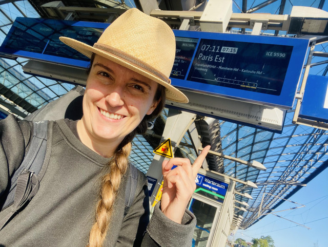 A person wearing a straw hat smiles and points at a departure board showing train information. The board indicates a train to Paris Est departing at 7:15. The background features a modern station with a glass roof.