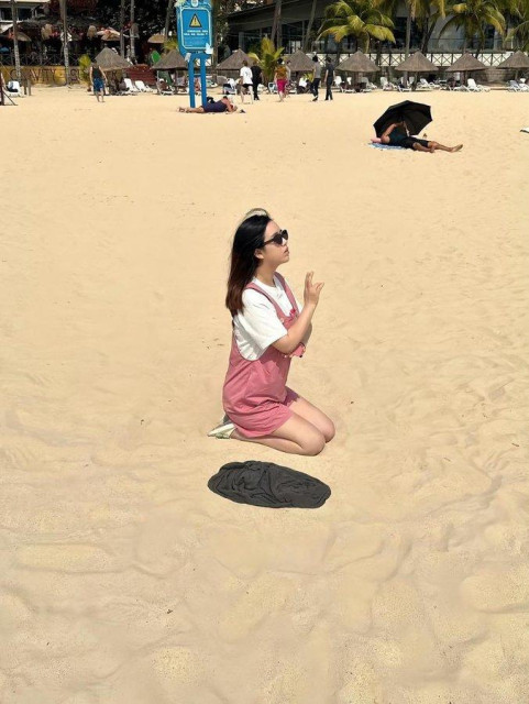 woman in white shirt and pink shorts is sitting on a beach. There's a piece of clothing next to her but the trompe l'oeil makes it looks like she is floating above a shadow. she also pretends to be praying. 