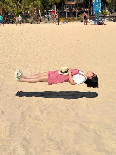 woman in white shirt and pink shorts is lying and resting on a beach. There's a piece of clothing next to her but the trompe l'oeil makes it looks like she is floating above a shadow.  She has a brown hat on her tummy. 