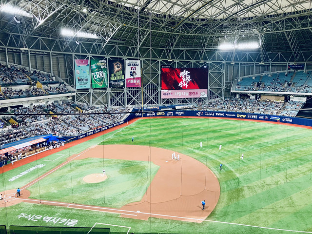 Wide-angle view from the seating area inside a domed baseball stadium during a game. The green field and brown infield are seen from behind home plate through protective netting. A pitcher stands on the mound while several infielders gather near second base during a pause in play, with an umpire and another staff member visible on the field. The large video board in center field displays a red graphic with Korean text, surrounded by advertising banners. Spectators fill much of the lower bowl, while many seats in the upper-right section remain empty. The steel roof trusses and bright stadium lights span the ceiling overhead.