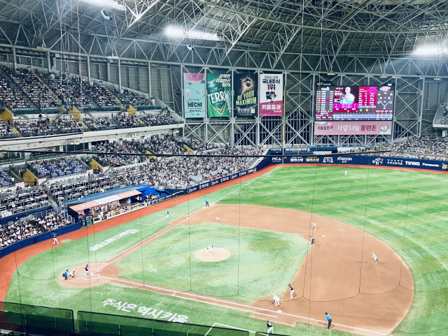 Wide-angle view of a baseball game inside a large domed stadium, photographed from high along the first-base side behind protective netting. The pitcher is delivering to a left-handed batter while runners are on first and second, with infielders positioned for the play. Thousands of spectators fill the lower and upper seating bowls. A large video scoreboard in right-center field displays the current batter and game information, surrounded by colorful Korean advertisements. The bright green artificial turf, brown infield, exposed steel roof trusses, and powerful stadium lighting emphasize the scale of the indoor ballpark.