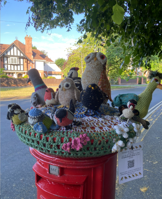 Knitted and crocheted group of birds topping a red postbox, with reference to NewlandParkBirds in a QR code