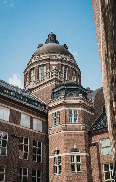 A dramatic low-angle view of the Swedish Museum of Natural History in Stockholm, showing its ornate brick facade and large copper-green dome topped by a small spire and weathervane, photographed against a clear blue summer sky. The building’s facade features tall arched and rectangular windows with light stone trim contrasting against the warm red-brown brick. Multiple rooflines and architectural tiers are visible. A narrow strip of an adjacent building’s wall is visible on the right edge of the frame. The composition emphasises height and verticality, with the dome dominating the upper portion of the image.