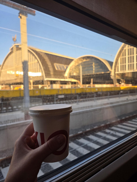 Holding a paper cup of coffee in front of a train window with the sunlit Amsterdam Central station in the background.