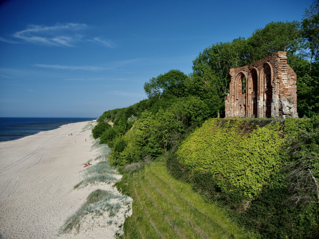 A picturesque beach scene featuring a sandy shoreline extending along the blue sea. In the foreground, there are remnants of a brick structure on a grassy cliff, surrounded by lush greenery. A few people are seen relaxing on the beach under a clear blue sky.