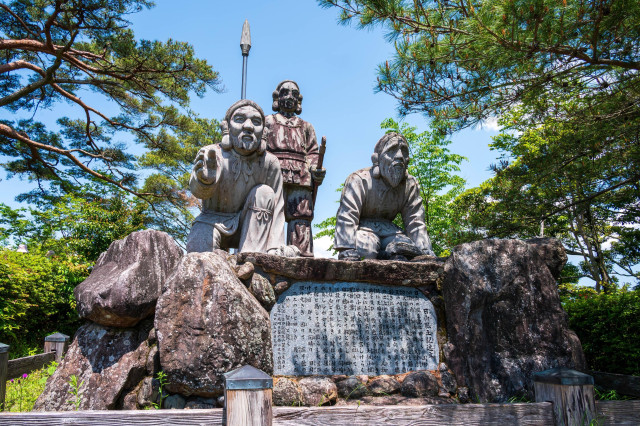 A sculpture depicting three figures representing indigenous people, set against a backdrop of trees and blue sky. In front of the figures is a stone plaque with inscriptions.