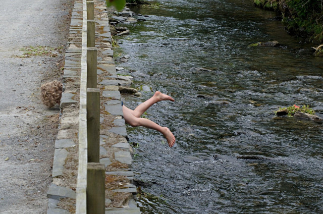 A photo showing a stone wall running next to a small river. one can see bare legs and curly hair of a person sticking out right and left from the wall, either trying to climb out of the river or into it through a hole in the wall.