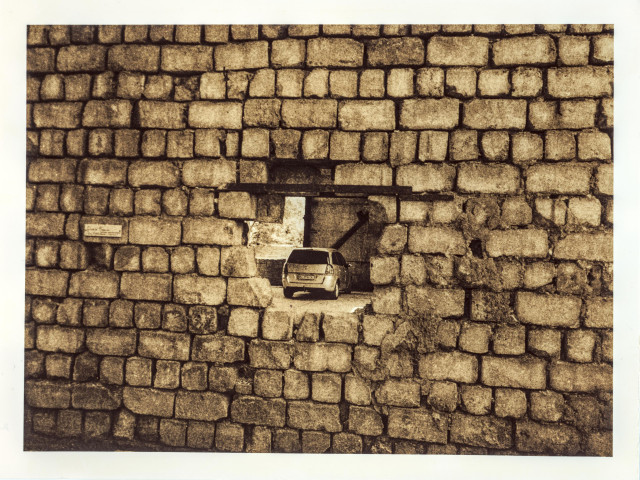 Darkroom lith print on Fomatone paper, 18×24 cm. A weathered brick wall fills the frame — the bricks worn and chipped with age, their edges crumbling in places.
Near the center, an opening breaks through the wall — its corners softened and rounded, unlike the brick pattern surrounding it. Through the gap, a covered parking area is visible, with a car parked inside. A small plaque is mounted on the wall to the left of the opening. The lith process renders the brick in warm, grainy sepia tones with deep, chalky blacks