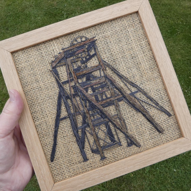 Stitched artwork depicting an old and rusted pit head winding wheel. Stitched on hessian, of which a lot is showing (negative space) and framed in a bare wood frame which tones with the fabric and subject. Held in the artist's hand for scale (8x8 inch frame)