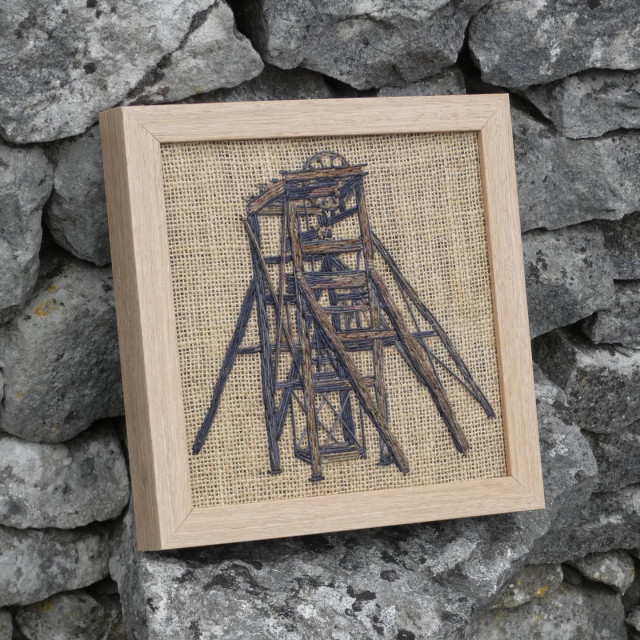 Same framed artwork leaning against a dry stone wall, viewed on an angle from the left