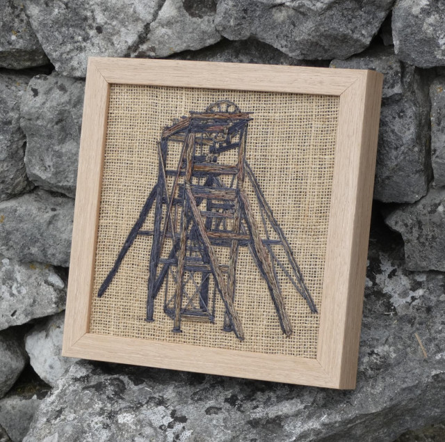 Same framed artwork leaning against a dry stone wall, viewed on an angle from the right
