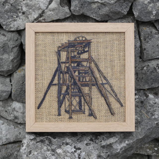 Same framed artwork leaning against a dry stone wall, viewed face on
