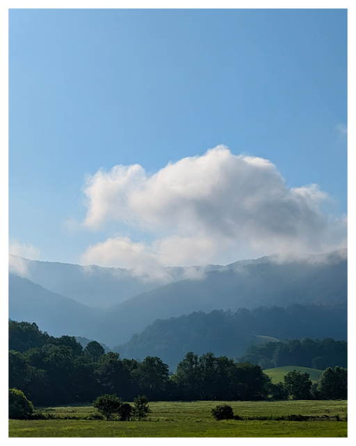 a scenic landscape with green fields in the foreground, densely wooded hills in the middle, and blue mountains in the background. fluffy white clouds hover over the hills under a clear blue sky,