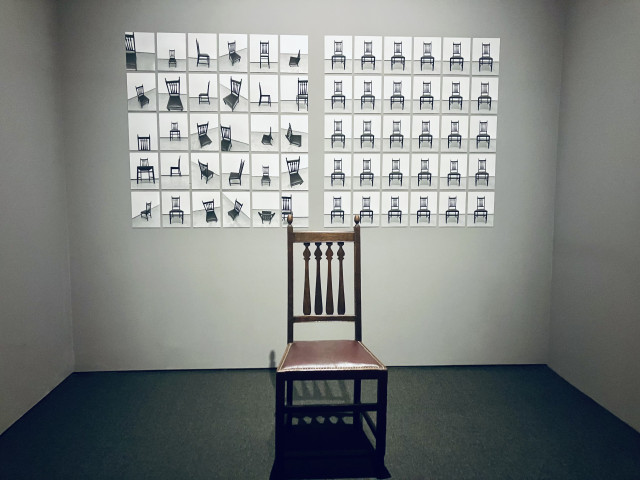 A wooden chair sits alone in the foreground of a sparsely lit gallery. On the wall behind it are two grids of small photographs of the same chair. The left grid shows the chair from many different angles and positions, while the right grid repeats a nearly identical front-facing view in each image. The minimalist installation emphasizes repetition, perspective, and the relationship between the physical chair and its photographic representations.