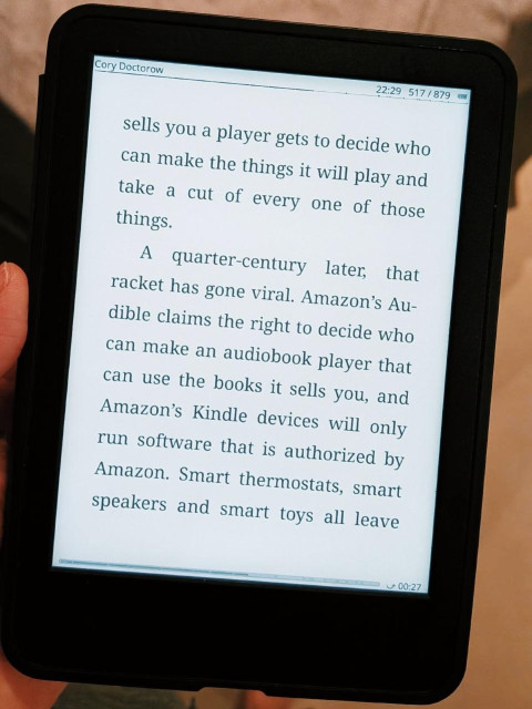 A photo of a hand holding a Kindle on a page that reads: “… sells you a player gets to decide who can make the things it will play and take a cut of every one of those things.

A quarter-century later, that racket has gone viral. Amazon's Audible claims the right to decide who can make an audiobook player that can use the books it sells you, and Amazon's Kindle devices will only run software that is authorized by Amazon. Smart thermostats, smart speakers and smart toys all leave…”