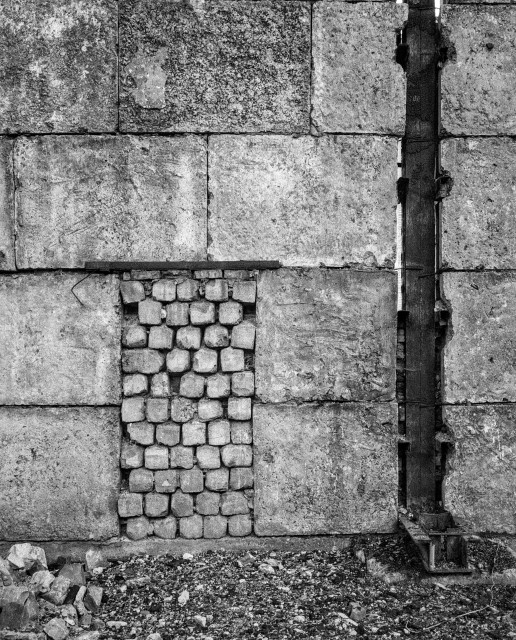 Black-and-white photograph of a weathered concrete block wall showing a former doorway between rooms that has since been bricked up, its weathered bricks distinct in texture from the surrounding concrete. A vertical rusted metal pipe runs down a seam on the right side of the frame, with loose rubble scattered along the base of the wall.