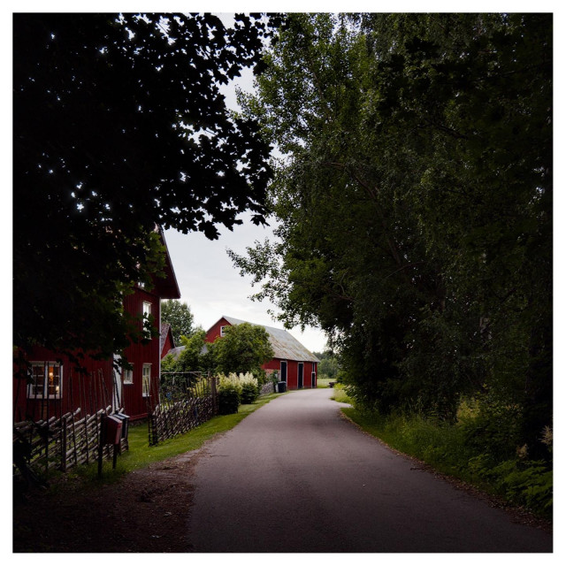 En fyrkantig bild på en asfalterad väg mellan träd och till vänster ligger ett rött trähus innanför ett staket av trä. Längre bort ligger en lada i rött trä med svarta dörrar.