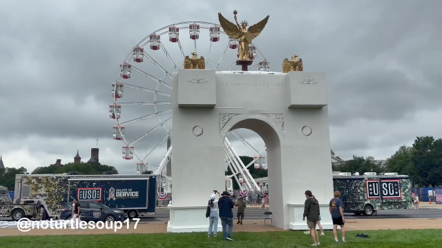  large white triumphal arch topped with a golden eagle statue with outstretched wings sits in front of a Ferris wheel under an overcast sky. The arch features the text "ONE NATION UNDER GOD" inscribed on its upper section. To the left and right of the structure are large dark trailers; the trailer on the left includes the text "USO" and "SALUTE TO SERVICE" amidst floral patterns, while the trailer on the right displays "USO". A group of people is gathered near the base of the arch in an open area with green grass in the foreground. The watermark "[@]noturtlesoup17" is visible in the bottom left corner.