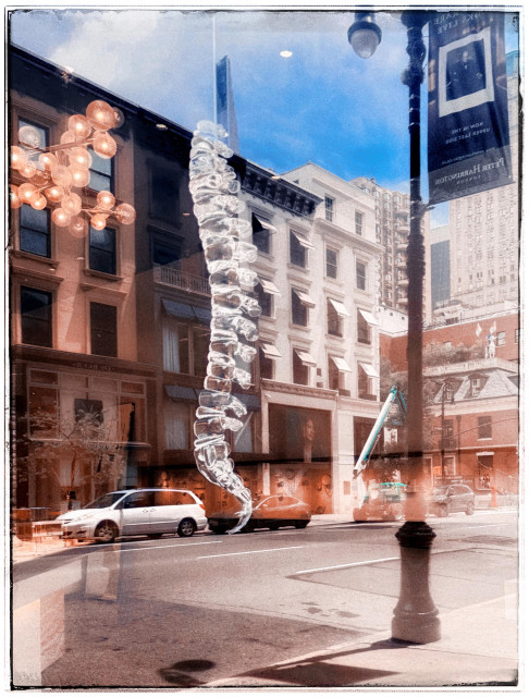 A color photo that was taken looking into the window of a interior decorating place. The reflection from the window is many brown stones across the street and a large lamp post with a banner on it, as well as a white car towards the bottom and the sidewalk. Coming through from inside the store is a light fixture on the right hand side that is made up of more than a dozen little round light orange bubbles with light inside of them. But the main focus of the photo is a glass vertebrae that was hanging in the window. It looks just like the skeleton of your spine only made out of glass