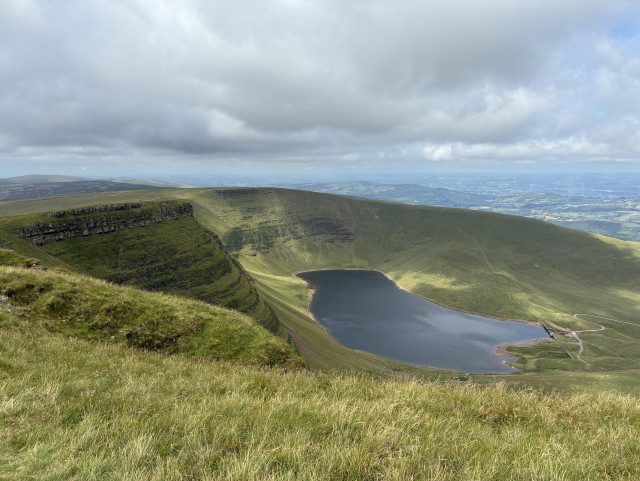 A photo from a mountain ridge cupping a lake in a cwm formed by a glacier. A steep, rock escarpment rises up above the lake on the left, the layers of rock softened by lush green vegetation. Long grass blown by the wind in the foreground. The sky is cloudy, though it was a short cloudy interlude in an otherwise sunny day. Countryside spreads out in the distance like a hazy green patchwork quilt.