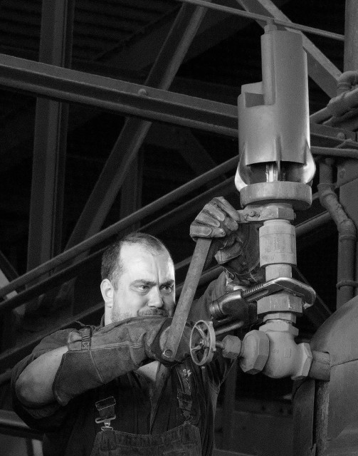 A black and white portrait photo of a man working on top of a steam engine. We see the top of the steam engine and the man's chest and shoulders as he stares intently toward the right on the large nut he is turning with a very large adjustable wrench. The nut holds the steam engine's whistle to the supply line. The metal roof rafters of the engine house are dimly lit in the background. 