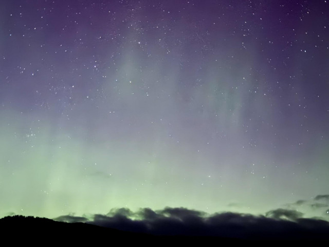 A night sky featuring a gradient of purple and green hues, illuminated by stars, with a silhouette of hills at the bottom and wispy clouds near the horizon.