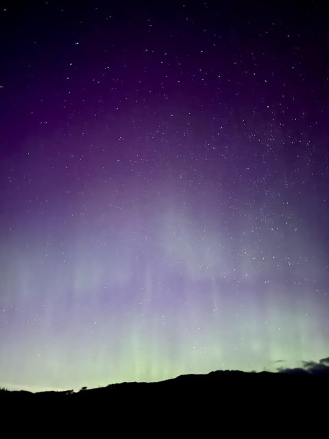 A night sky filled with stars, showcasing a gradient of purple and green hues, with a silhouette of a mountain range at the bottom.