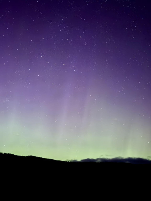 A night sky filled with stars, featuring a gradient of purple and green hues near the horizon, and silhouetted hills below.